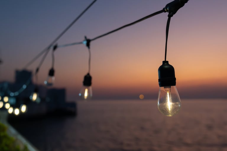 Decorative string light bulbs are hanging above calm purple sea water with sunset in evening