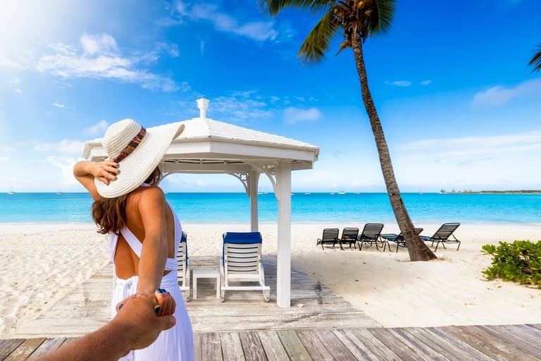 Woman in white hat and sundress relaxing on tropical beach with white pavilion, lounge chairs, and palm trees overlooking turquoise ocean