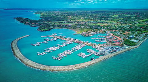 Aerial view of a coastal marina with white boats docked in turquoise waters protected by a curved breakwater