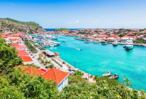 Aerial view of a colorful tropical harbor with turquoise water, red-roofed buildings, boats, and lush vegetation surrounding the bay