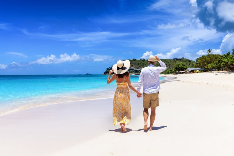 Couple walking on pristine white sand beach holding hands, wearing sun hats, with turquoise ocean and blue sky in background